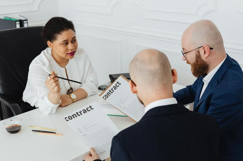 Three business professionals in formal attire discussing a contract in a modern office setting